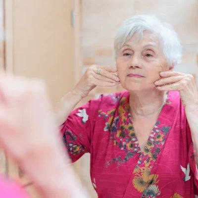 Femme âgée appliquant un soin sur son visage devant un miroir, pour illustrer les routines naturelles visant à prendre soin de la peau mature.